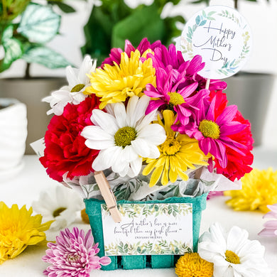 Green berry basket holding flowers. Has a round tag sticking out of the flowers that says Happy Mother's Day. Also has a rectangular tag clipped to the front of the basket with a mini clothespin. The tag reads: 